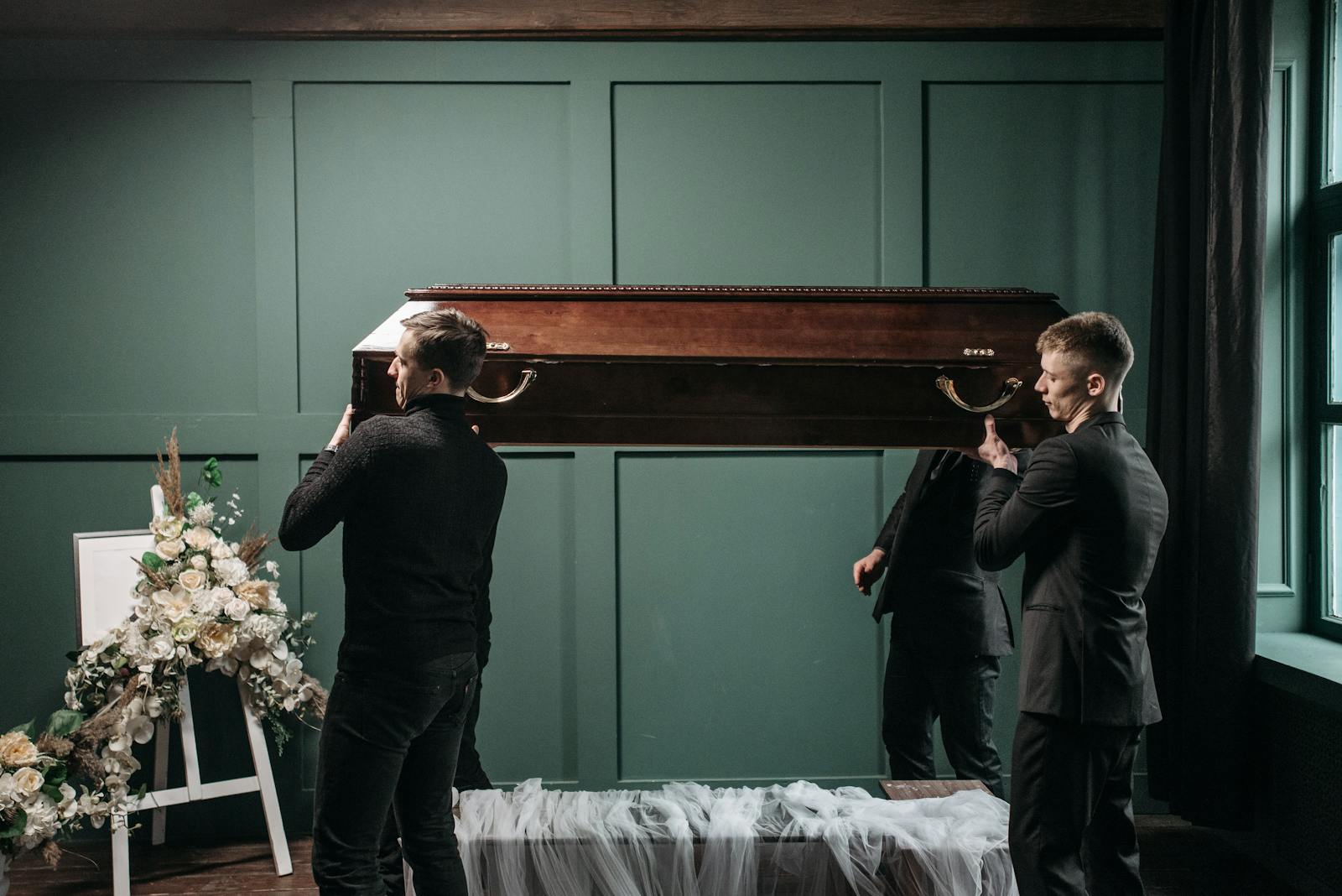 Pallbearers carry a wooden coffin at an indoors funeral service, surrounded by floral tributes.